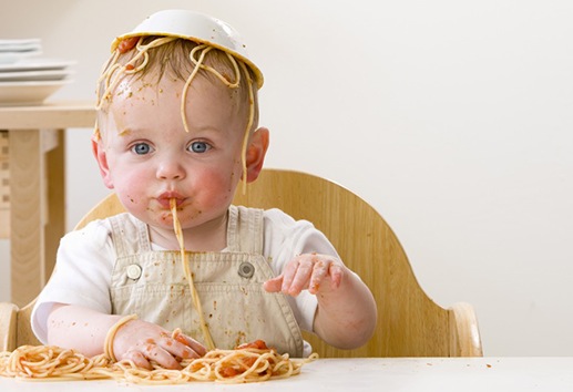 Messy baby boy in high chair with bowl of spaghetti on head