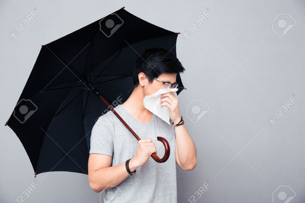 39439599-Sick-asian-man-holding-umbrella-and-blowing-nose-over-gray-background-Stock-Photo